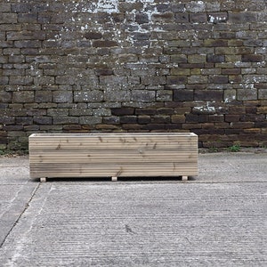 May include: A long, rectangular wooden planter box with a natural wood finish. The planter is sitting on a concrete surface in front of a brick wall.
