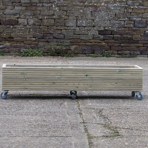 May include: A wooden planter box on wheels. The planter is made of light brown wood and has a rectangular shape. The wheels are black with blue rims.