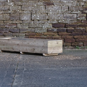 May include: A long, rectangular wooden planter box sits on a grey concrete surface in front of a brick wall. The planter is made of natural wood and has a simple, rustic design.