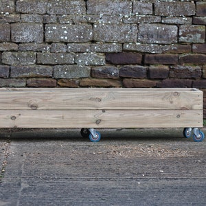 May include: A wooden planter box on wheels. The planter is made of natural wood and has a light brown finish. The wheels are black with blue rubber tyres.
