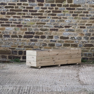 May include: A long, rectangular wooden planter box with a natural wood finish. The planter is sitting on a concrete surface in front of a stone wall.
