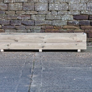 May include: A long, rectangular wooden planter box with a natural finish. The planter is sitting on a concrete surface in front of a brick wall.