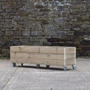 May include: A long, rectangular wooden planter box on wheels. The planter is made of natural wood and has a rustic look. It is sitting on a concrete surface in front of a stone wall.