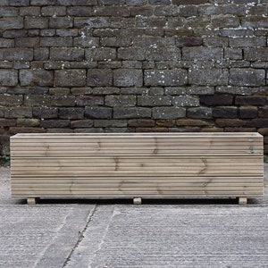 May include: A long, rectangular wooden planter box with a slatted design. The planter is made of natural wood and has a rustic look. It is sitting on a concrete surface in front of a brick wall.