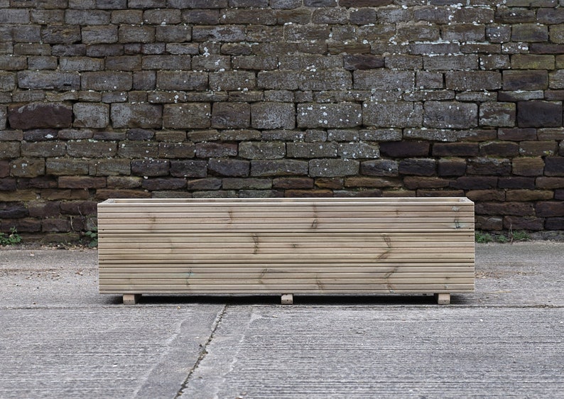 May include: A long, rectangular wooden planter box with a slatted design. The planter is made of natural wood and has a rustic look. It is sitting on a concrete surface in front of a brick wall.