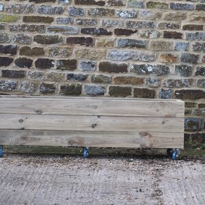 May include: A wooden planter box on wheels, made of natural wood with a light brown finish. The planter is sitting in front of a stone wall.