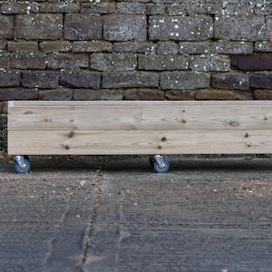 May include: A wooden planter box with wheels, made of light brown wood. The planter is sitting on a grey concrete surface in front of a brick wall.