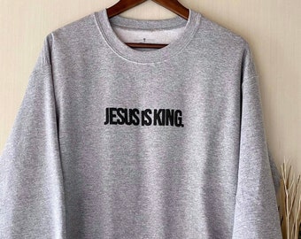 以下が含まれることがあります： 前面に黒い文字で「JESUS IS KING.」とプリントされた灰色のスウェットシャツです。