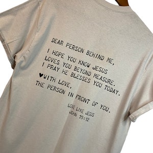 Op de afbeelding: Een beige T-shirt met een boodschap op de achterkant. De boodschap luidt: "Lieve persoon achter me, ik hoop dat je weet dat Jezus van je houdt boven alle maat. Ik bid dat hij je vandaag zegent. Met liefde, de persoon voor je. Lief zoals Jezus Johannes 15:12"