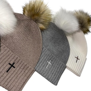 May include: Four knit beanies in neutral colors: taupe, gray, white, and cream. Each beanie features a faux fur pom-pom and a cross embroidered on the front. The beanies are a winter accessory.
