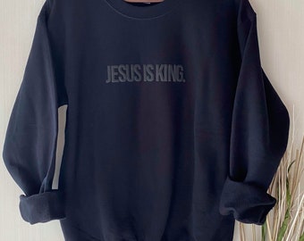 以下が含まれることがあります： 胸に「JESUS IS KING」の文字が同系色でプリントされた黒いスウェットシャツ。木製のハンガーに掛けられており、袖は折り返されている。背景はニュートラルカラー。
