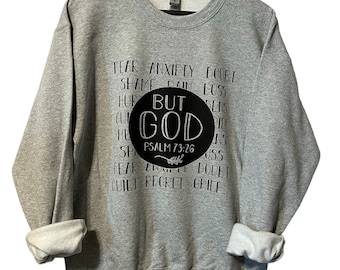 以下が含まれることがあります： 黒い円の中に「But God」と書かれたグレーのスウェットシャツ。その下に聖書の引用「詩篇73:26」が書かれている。円は「恐怖」「不安」「疑い」「恥」「痛み」「損失」「傷つき」「罪悪感」「後悔」「悲しみ」などの言葉で囲まれている。