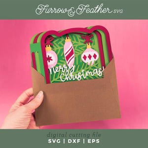 May include: A layered Christmas card with three ornaments and the words "Merry Christmas" in white. The card is inside a brown envelope, held against a pink background. The image includes the text "Furrow & Feather SVG" and "digital cutting file SVG | DXF | EPS".
