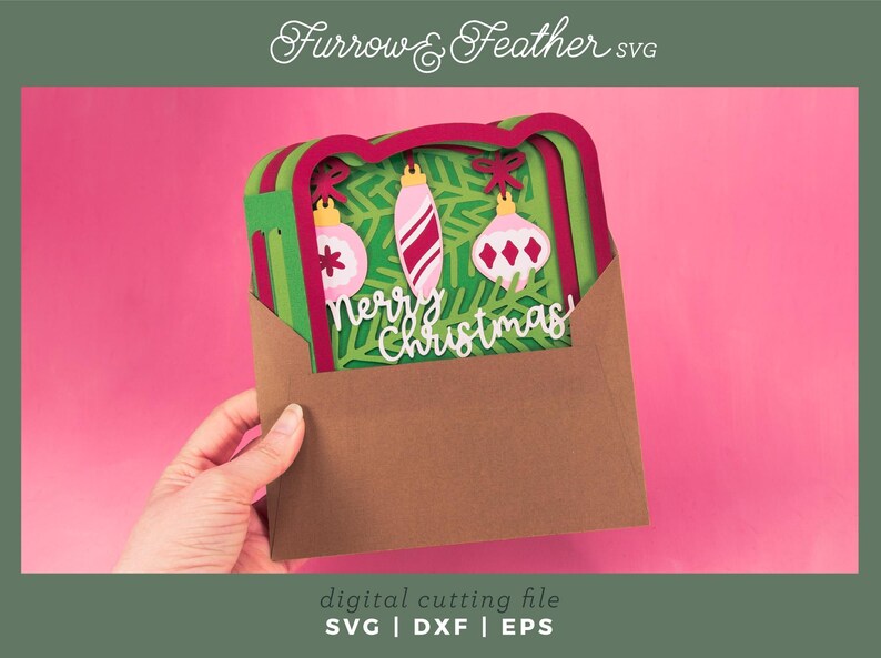 May include: A layered Christmas card with three ornaments and the words "Merry Christmas" in white. The card is inside a brown envelope, held against a pink background. The image includes the text "Furrow & Feather SVG" and "digital cutting file SVG | DXF | EPS".