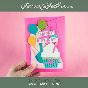 May include: A pink card with a pop-up cupcake design. The cupcake is white with pink frosting and a green and blue wrapper. The card has a banner that says "Happy Birthday!" with balloons in the background.