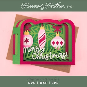 May include: A layered Christmas card with a pink and green design. The card features three hanging ornaments and the words "Merry Christmas". The card is set against a pink background.