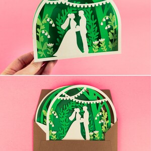 May include: A green and white layered paper cut design of a wedding couple standing under a canopy of trees with string lights. The design is in a brown envelope.