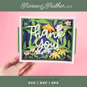 May include: A paper cut-out card with a floral design and the words "Thank You". The card features green leaves, colorful flowers, and a dark blue background. The card is held against a pink background.
