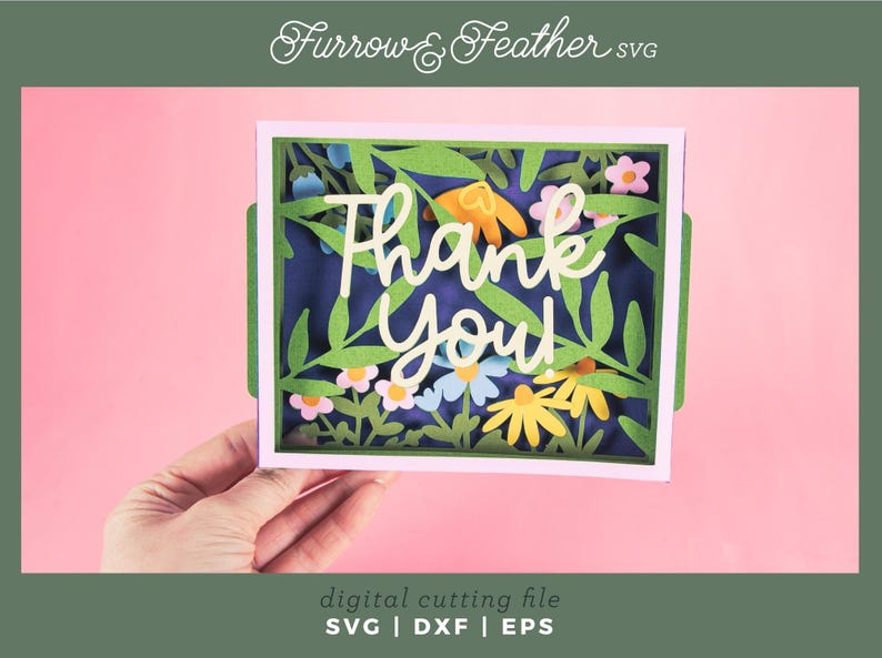 May include: A paper cut-out card with a floral design and the words "Thank You". The card features green leaves, colorful flowers, and a dark blue background. The card is held against a pink background.