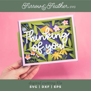 May include: A papercraft card with the words "Thinking of you" in white script. The card features a floral design with green leaves and colourful flowers. The background is a deep purple, and the card is held against a pink background.