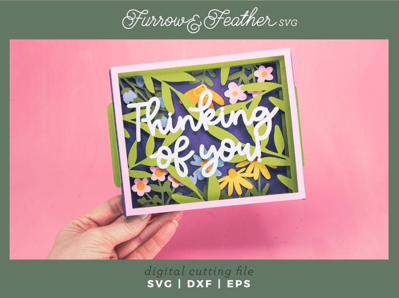 May include: A papercraft card with the words "Thinking of you" in white script. The card features a floral design with green leaves and colourful flowers. The background is a deep purple, and the card is held against a pink background.