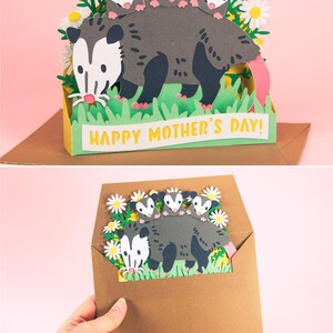 May include: A papercraft Mother's Day card depicting a grey opossum with three babies, white daisies, and green grass. The card reads "Happy Mother's Day!" in yellow. The card is presented inside a brown envelope, perfect for gifting.