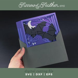 Pode incluir: Uma pe&ccedil;a de arte em papel em camadas com tema de Halloween. O design inclui morcegos pretos, uma lua crescente branca e &aacute;rvores estilizadas em um fundo roxo. A obra de arte &eacute; mantida em um envelope cinza. O texto na imagem diz "Furrow & Feather SVG" e "digital cutting file".