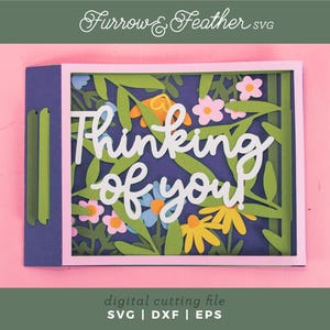 May include: A paper card with the words "Thinking of you" in white script, featuring a floral design with green leaves and colourful flowers. The background is a deep blue, set against a pink backdrop. The card is a digital cutting file.