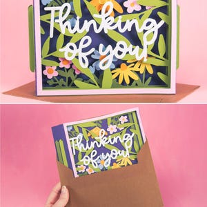 May include: A paper art card with the words "Thinking of you" in white script. The card has a floral design with green leaves, yellow and pink flowers, and a purple background. The card is in a brown envelope.
