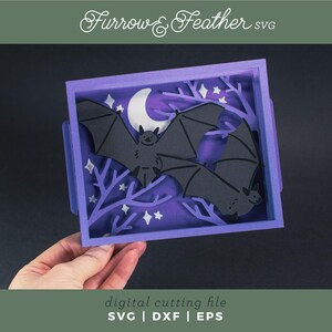 Pode incluir: Uma pe&ccedil;a de arte em papel em camadas com dois morcegos pretos em silhueta contra um fundo roxo com uma lua crescente e estrelas. A obra de arte est&aacute; em uma moldura roxa com o texto "Furrow & Feather SVG" e "digital cutting file SVG | DXF | EPS".