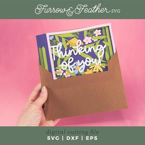 May include: A papercraft card with the words "Thinking of you" in white script, surrounded by colourful paper flowers and foliage. The card is inside a brown envelope, set against a pink background. The image also includes the text "digital cutting file SVG | DXF | EPS".