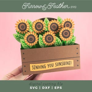 May include: A papercraft of sunflowers in a brown wooden crate with the text "SENDING YOU SUNSHINE!". The sunflowers have yellow petals and brown centres, with green leaves. The image is a digital cutting file.