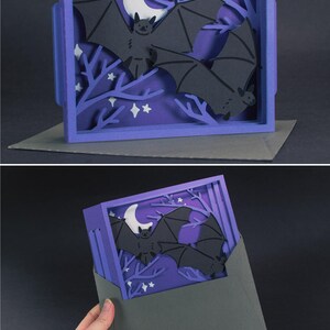 Pode incluir: Um cart&atilde;o de arte em papel em camadas com tema de Halloween. O cart&atilde;o tem uma moldura roxa com uma cena noturna, incluindo morcegos, uma lua crescente, estrelas e galhos de &aacute;rvores. O cart&atilde;o est&aacute; em um envelope.