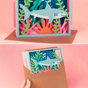 Puede incluir: Una pieza de arte en papel en capas que presenta un tibur&oacute;n azul claro nadando en una escena submarina con coral y algas marinas. La obra de arte se muestra en un marco de caja con detalles en rosa y est&aacute; sobre una superficie marr&oacute;n. El texto "FURROW AND FEATHER SVG" est&aacute; en la parte inferior.