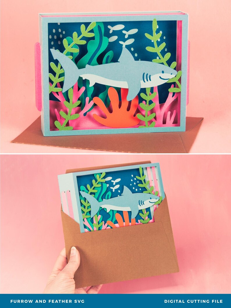 Puede incluir: Una pieza de arte en papel en capas que presenta un tibur&oacute;n azul claro nadando en una escena submarina con coral y algas marinas. La obra de arte se muestra en un marco de caja con detalles en rosa y est&aacute; sobre una superficie marr&oacute;n. El texto "FURROW AND FEATHER SVG" est&aacute; en la parte inferior.