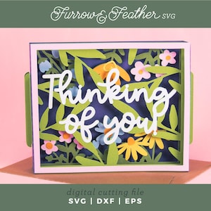 May include: A layered paper art card with a floral design and the words "Thinking of you." The card features green leaves, yellow and pink flowers, and a dark blue background. The card is in a white frame.