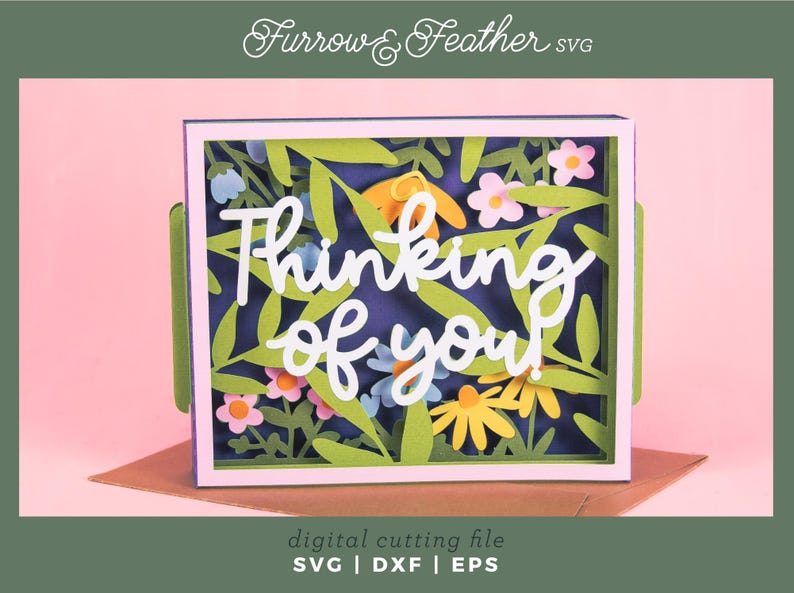 May include: A layered paper art card with a floral design and the words "Thinking of you." The card features green leaves, yellow and pink flowers, and a dark blue background. The card is in a white frame.