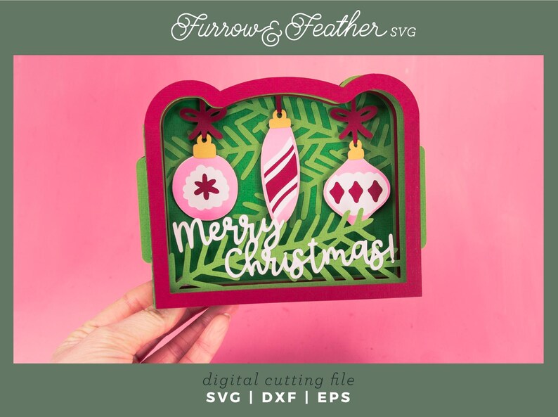 May include: A layered Christmas card design with a red frame, green accents, and pink background. The card features three hanging ornaments and the words "Merry Christmas!" in white. The design is a digital cutting file.
