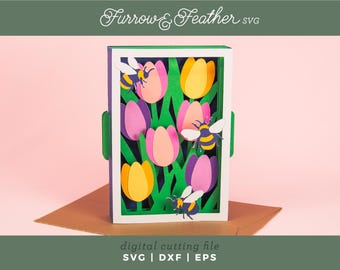 Könnte beinhalten: Ein Papier-Schattenkasten mit rosa und gelben Tulpen, grünem Laub und Bienen. Das Design ist vor einem rosa Hintergrund platziert, mit dem Text "Furrow & Feather SVG" oben. Der Text "digital cutting file" steht unten.