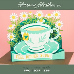 May include: A papercraft teacup and saucer with a tea bag, surrounded by daisies. The teacup is white with a teal rim and handle, sitting on a teal saucer. The words "FEEL BETTER SOON!" are displayed on a teal banner.