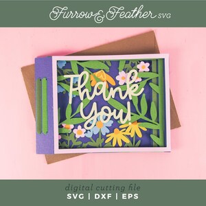 May include: A layered paper card with a floral design and the words "Thank You" in white script. The card features green leaves, yellow and white flowers, and a purple border. The background is a light pink color.
