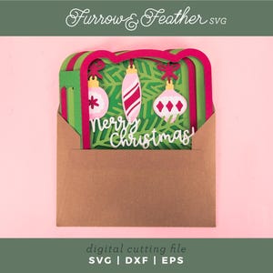 May include: A layered paper Christmas card design in a brown envelope. The card features green and red layers with three hanging ornaments and the words "Merry Christmas". The design is set against a pink background. The text "Furrow & Feather SVG" is at the top.