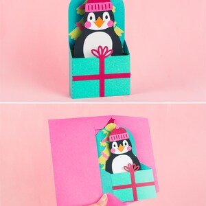 May include: A pop-up card featuring a penguin wearing a pink hat and sitting in a gift box. The penguin is in front of a green Christmas tree. The card is open and the penguin is popping up.