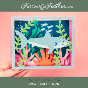 Puede incluir: Una escena de manualidades en papel con un tibur&oacute;n azul claro nadando en un entorno oce&aacute;nico. El dise&ntilde;o incluye algas verdes, coral naranja y peque&ntilde;os peces blancos sobre un fondo azul oscuro. El texto "Furrow & Feather SVG" est&aacute; en la parte superior.