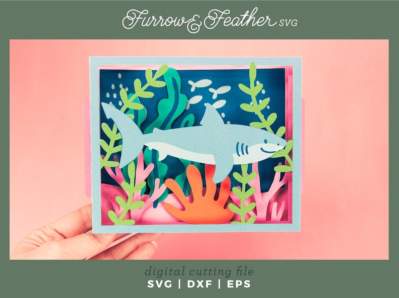 Puede incluir: Una escena de manualidades en papel con un tibur&oacute;n azul claro nadando en un entorno oce&aacute;nico. El dise&ntilde;o incluye algas verdes, coral naranja y peque&ntilde;os peces blancos sobre un fondo azul oscuro. El texto "Furrow & Feather SVG" est&aacute; en la parte superior.
