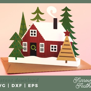 May include: A papercraft Christmas scene with a red cabin, white snow-covered roof, and green trees. A brown Christmas tree with a red star is in front of the cabin. The words "instant download" and "SVG | DXF | EPS" are at the bottom.