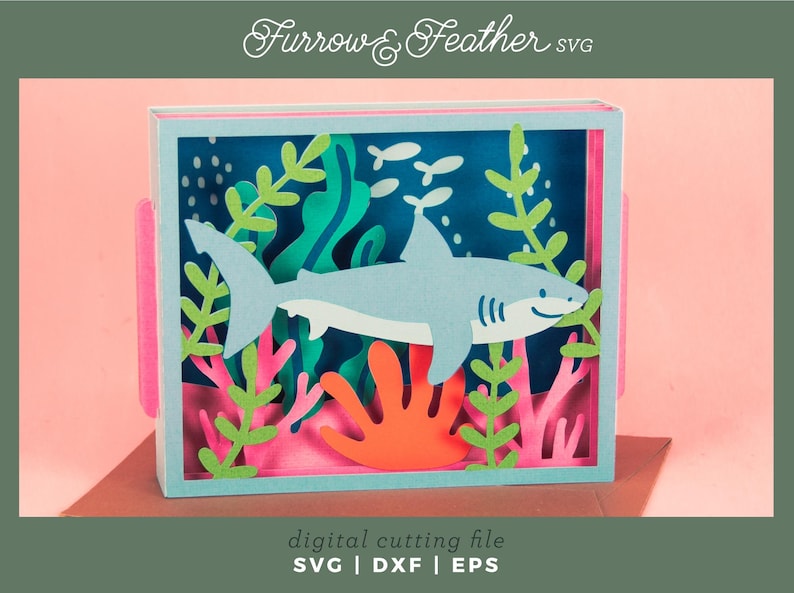 Puede incluir: Una obra de arte en papel en capas que representa un tibur&oacute;n azul claro nadando en una escena submarina. La escena incluye algas verdes, coral rosa y peque&ntilde;os peces blancos sobre un fondo azul oscuro. El texto "Furrow & Feather SVG" est&aacute; en la parte superior.