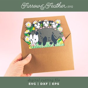 May include: A brown paper envelope contains a paper cut-out scene of a possum family amongst daisies. The possum is grey with a white face and pink tail. The text "Furrow & Feather SVG" is at the top.