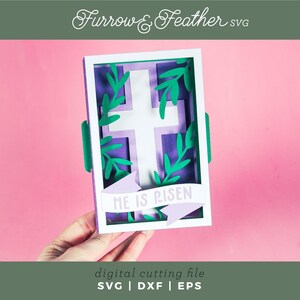 May include: A purple and white cross with green leaves surrounding it, inside a white frame. The frame has a purple banner with the text "HE IS RISEN" in white.