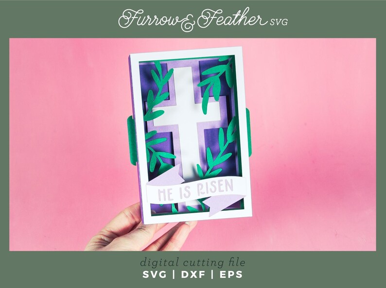 May include: A purple and white cross with green leaves surrounding it, inside a white frame. The frame has a purple banner with the text "HE IS RISEN" in white.